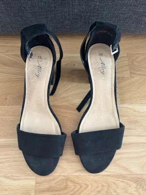 Svarta klackar  -  svarta pumps från Alley med öppen tå och bred rem över foten. Klackarna har en justerbar ankelrem med silverfärgat spänne och en mjuk innersula i beige. 
