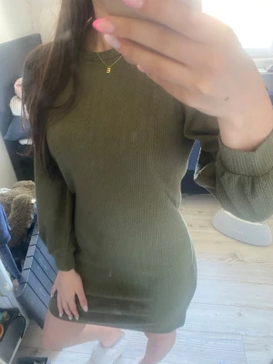 Olivgrön ribbad stickad klänning - Snygg olivgrön stickad klänning med ribbad struktur och långärmad design. Kort modell med rund halsringning och en kroppsnära passform som framhäver figuren. Perfekt för dig som gillar enkel men trendig stil.