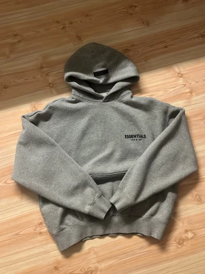 Grå Essentials hoodie - Snygg grå hoodie från Essentials med svart tryck på bröstet där det står 'ESSENTIALS Fear of God'. Klassisk passform med stor huva och magficka. Tillverkad i mjuk bomullsblandning, perfekt för en avslappnad streetwear-look.