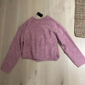 Ljusrosa stickad tröja 157 - Jättesnygg ljusrosa stickad tröja från 157 med rund halsringning och långa ärmar. Tröjan har en avslappnad passform och ribbstickade muddar vid ärmslut och nederkant. Perfekt för lager på lager och en chill look.