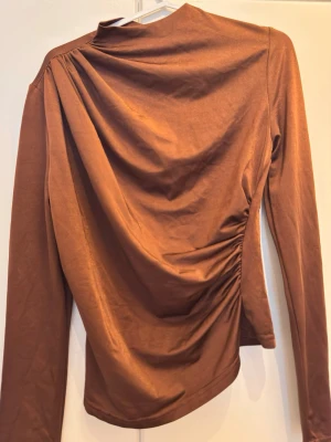 Brun långärmad topp med snörning - Snygg brun långärmad topp med hög hals och draperad snörning i sidan som ger en cool asymmetrisk look. Tillverkad i mjukt material med en slim passform som framhäver siluetten. Perfekt för dig som vill ha något trendigt och stilrent.