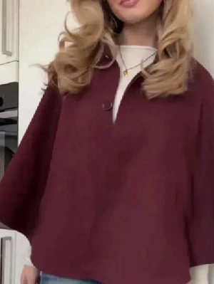 Vinröd cape-kappa med knapp - Stilren vinröd cape-kappa med lös passform och en dekorativ knapp framtill. Kappan har en elegant, vid siluett och är perfekt för lager-på-lager. Materialet ser ut att vara mjukt och något tjockare, vilket ger en lyxig känsla.