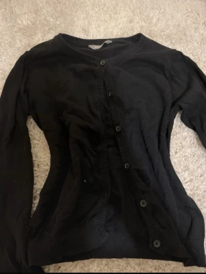 Svart fin kofta med knappar - En enkel svart kofta med rund halsringning och knäppning framtill. Långärmad modell med ribbade muddar vid ärmslut och nederkant. Perfekt att slänga över en t-shirt eller linne för en clean look. Storlek 146/152 men sitter som en xxs.