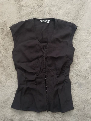 Svart korsett topp från Weekday  - Snygg svart korsett topp från Weekday med djup V-ringning och hyskknäppning framtill. Figurnära passform och ärmlös design ger en modern och edgy look. Tillverkad i ett mjukt tyg som känns bekvämt mot huden. Perfekt att styla med jeans eller kjol.