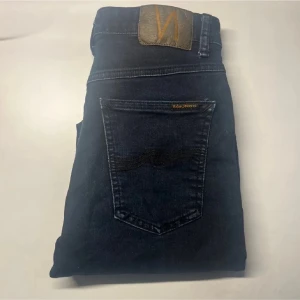Nudie jeans  - Snygga mörkblå jeans från Nudie Jeans i slim fit. Klassisk femficksmodell med guldfärgade detaljer och Nudie-logga på bakfickan. Jeansen har hög midja och är tillverkade i stretchigt denim för en bekväm passform. Modellapp saknas. Midja 34cm, längd 99cm.  Ifall det inte finns någon bild i annonsen där jag bär plaggen jag säljer så är det för att jag ej kan ha storleken heller passformen, så kolla måtten för storlek och googla på modellnamnet för passform 😉 s3