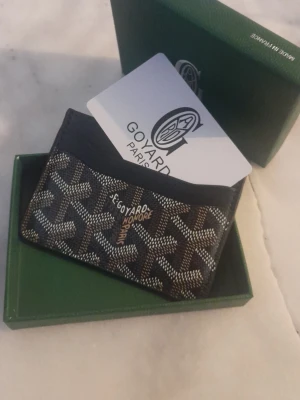 Svart kortfodral  - Snyggt svart kortfodral med det klassiska mönstret i vitt och brunt. Fodralet har flera fack för kort med läderdetaljer. Perfekt för dig som vill ha en stilren och exklusiv accessoar.