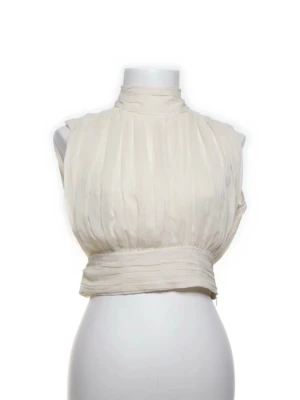 Vit omlott blus med hög krage - Säljer en elegant vit blus med hög krage och omlott-detalj i midjan. Blusen är ärmlös och har ett lätt transparent, plisserat tyg som ger en luftig känsla. Perfekt för dig som gillar stilrena och trendiga toppar.