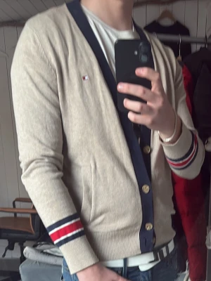 Beige cardigan från Tommy Hilfiger - Snygg beige cardigan från Tommy Hilfiger med marinblå knappslå och muddar med röd, vit och marinblå rand. Liten logga på bröstet och två fickor framtill. Perfekt för lager på lager och enkel att matcha med jeans eller t-shirt.