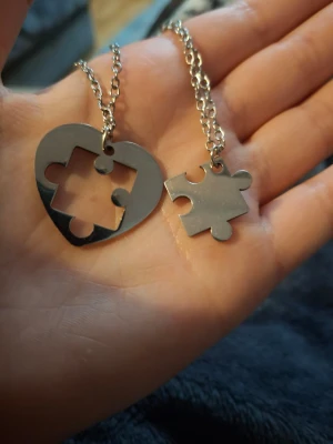 Halsband med hjärta och pusselbit - Två matchande halsband i silverfärgad metall, ett med ett hjärta med utskuren pusselbit och ett med en pusselbitsberlock. Perfekt för att dela med någon speciell. Kedjorna är tunna och smidiga, och designen är modern och stilren.