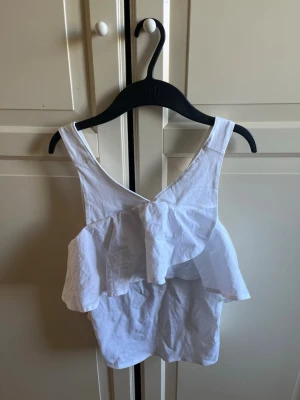 Vit volang topp med breda axelband - Söt vit topp med breda axelband och dubbla volanger framtill. Toppen har en luftig och somrig känsla, perfekt för varma dagar. Materialet känns som bomull och modellen är ärmlös med en lätt croppad passform.