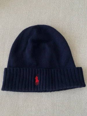 Mörkblå mössa från Polo Ralph Lauren - Snygg mörkblå ribbstickad mössa från Polo Ralph Lauren med klassisk röd broderad logga framtill. Tillverkad i mjuk merino-ull som håller dig varm. Perfekt accessoar för kyliga dagar och enkel att matcha med olika stilar.