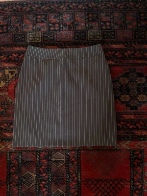 Brun kortkjol med vita ränder - Snygg brun kortkjol med tunna vita vertikala ränder. Kjolen har en klassisk, rak passform och stängs med dragkedja bak. Perfekt för en stilren och trendig look.