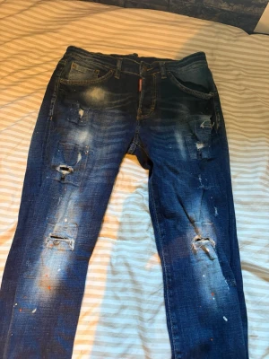 Blå dsquared2 jeans - Säljer ett par blå jeans med slitningar och hål på benen samt coola färgstänk för en trendig och avslappnad look. Jeansen har fem fickor, normal midja och raka ben. Perfekta för dig som gillar streetstyle och vill sticka ut lite extra.