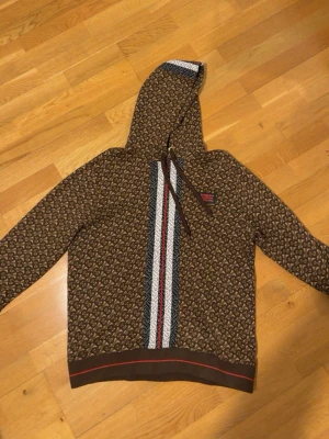 Brun Burberry hoodie med monogram (Äkta) (L passar M) - Snygg brun hoodie från Burberry med ikoniskt TB-monogram över hela plagget. Framtill finns en randig detalj i vitt, svart och rött samt en patch med 'Burberry London England'. Hoodien har dragsko i huvan och ribbade muddar. Perfekt för dig som gillar streetstyle och designerplagg.