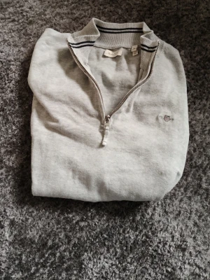 Ljusgrå halvzip GANT - Ljusgrå långärmad tröja med halv dragkedja och diskret broderad logga på bröstet. Tröjan har ribbade muddar vid ärmslut och nederkant samt randig detalj i kragen. Perfekt för en clean och stilren look.