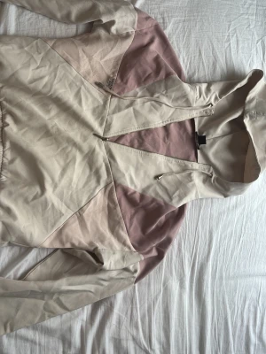 Beige och rosa hoodie från New yorker - Säljer en snygg hoodie från new yorker i beige och ljusrosa färgblock. Hoodien har dragkedja framtill, huva med snören och är tillverkad i mjukt bomullsmaterial. Perfekt för en chill och avslappnad stil.