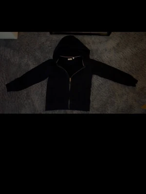 Gant zip hoodie - Klassisk svart hoodie från GANT med hel dragkedja framtill och huva. Insidan har mjuk fleece och kragen har en snygg detalj med röd och vit rand. Perfekt för en avslappnad och stilren look.