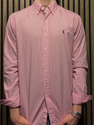 Rödvit randig skjorta från Ralph Lauren - Klassisk rödvit randig skjorta från Ralph Lauren med button-down krage och blå broderad logga på bröstet. 