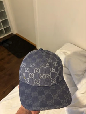 Blå Gucci keps med GG-mönster - Snygg blå keps från Gucci i denim-look med det klassiska GG-mönstret över hela kepsen. Böjd skärm och justerbar baksida. Perfekt för dig som vill ha en exklusiv och trendig accessoar till din outfit.