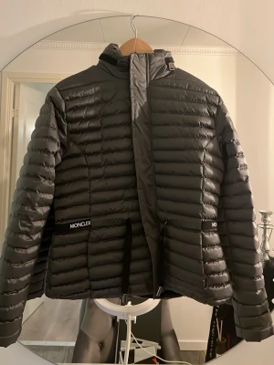 Svart dunjacka från Moncler med huva - Snygg svart dunjacka från Moncler med quiltad design och huva. Jackan har dragkedja framtill, två fickor med Moncler-logga och en lätt glansig finish. Perfekt för kalla vinterdagar och riktigt stilren look.