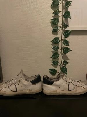 Philippe modells  - Säljer ett par vita sneakers med svarta detaljer på hälen och sidan. Skorna har klassisk låg profil och snörning framtill. Materialet är en mix av läder och textil, vilket ger en skön och stilren look. Opps visa detaljer ska va på skon det är designen 