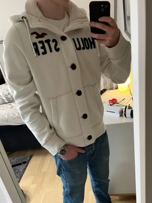 Vintage Hollister cardigan - Säljer nu denna snygga vita vintage Herr Hollister cardigan. Tröjan har blå Hollister text broderat på framsidan, samt en vinröd Hollister logga. Står att tröjan är i storlek XL, men passar som L/M.  Tröjan är nästan i ny skick och har inga defekter. Modellen på bilden är 183 och väger ca 68 kilo.  Längd: 68 cm Bredd (armhåla till armhåla): 58 cm Skriv gärna om ni har några mer funderingar🙂