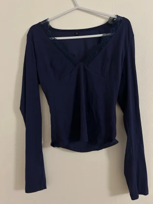 Mörkblå långärmad topp med spets - Mörkblå långärmad topp med v-ringning och spetsdetaljer vid halsen. Toppen har en figurnära passform och är tillverkad i ett mjukt, stretchigt material. Perfekt för dig som gillar stilrena plagg med en liten twist.