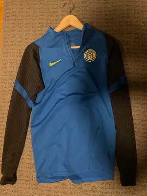 Blå Inter Nike träningströja - Snygg blå träningströja från Nike med Inter-logga på bröstet och gul Nike-swoosh. Tröjan har svarta långärmade ärmar med diskret mönster och en half zip-dragkedja vid halsen. Perfekt för fotbollsträning eller sportiga dagar.