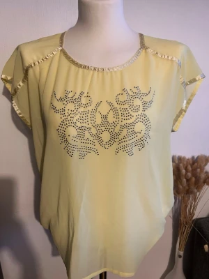 Gul topp med paljettdetaljer - Snygg gul topp med korta ärmar och glittriga paljettdetaljer längs halsringning och ärmslut. Framsidan har ett dekorativt mönster av små silverfärgade nitar. Luftigt material som passar perfekt till varma dagar.