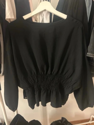 Svart blus med smockad midja - Trendig svart blus med rund halsringning och smockad midja som ger en snygg siluett. Blusen har långa ärmar med lätt puff och är tillverkad i ett tunt, mjukt tyg som faller fint. Perfekt att styla med både jeans och kjol.