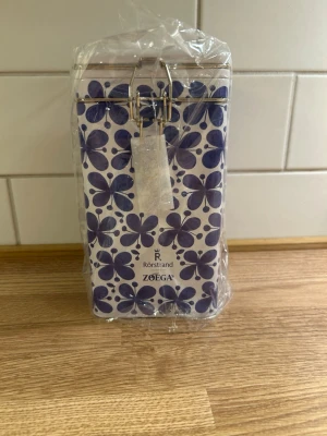 Blommig plåtburk Rörstrand Zoégas - Säljer en snygg plåtburk från Rörstrand i samarbete med Zoégas. Burken är vit med blå-lila blommönster och har ett metallspänne för förslutning. Perfekt för kaffe eller småförvaring. Materialet är metall och designen är riktigt stilren.