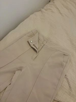 Beige kostymbyxa med pressveck - Snygga beige kostymbyxor med raka ben och markerade pressveck framtill. Byxorna har hög midja, diskreta sidofickor och stängs med dragkedja och hake. Perfekta för en clean och stilren look. Passade mig inte i storlek och därför aldrig använts