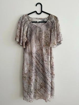 Branting klänning - Med Innerklänning - Strl S - Snygg och luftig klänning från Branting i ett mjukt beige/brunt mönster. Klänningen har en lös och bekväm passform med fina volangärmar och knytning framtill vid halsen.  Den har en beige innerklänning som gör att den inte är genomskinlig och faller väldigt fint.  Märke: Branting Storlek: S Längd: ca 88 cm