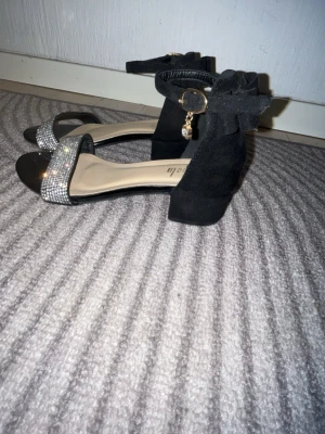 Svarta pumps med strass och rosett - Säljer ett par svarta pumps med bred klack, dekorerade med strass framtill och en stor rosett bak på hälen. Skorna har ankelrem med guldfärgad detalj och liten hängande berlock. Materialet ser ut som mocka och innersulan är ljus. Perfekta för att sticka ut.