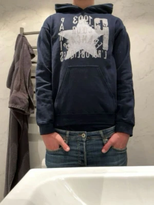 Mörkblå hoodie med tryck - Mörkblå hoodie med vit grafisk text och stjärntryck på bröstet. Klassisk känguruficka framtill och huva. Perfekt för en avslappnad stil och enkel att matcha med jeans.