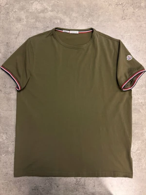 Olivgrön Moncler t-shirt med logga - Snygg olivgrön t-shirt från Moncler med korta ärmar och rund hals. Ärmsluten har en röd, vit och blå randig detalj och Moncler-loggan sitter på vänster ärm. Perfekt för en clean och stilren look.
