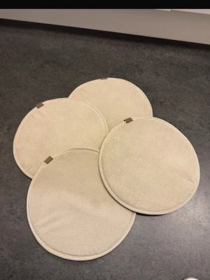Beige runda sittdynor 4-pack - Fyra stycken runda sittdynor i beige tyg med små bruna hängöglor. Perfekta att lägga på stolar för extra komfort. Passar bra i köket eller på uteplatsen. Tygmaterial och enkel, stilren design.