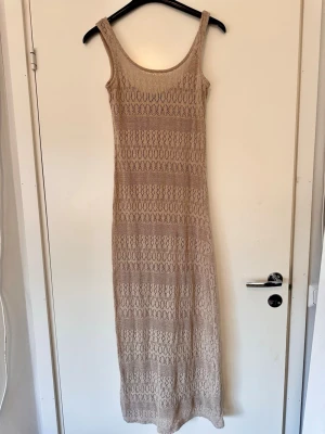Beige stickad strandklänning  - Snygg beige långklänning i stickat material med breda axelband och rundad ringning. Klänningen har ett genomgående hålmönster som ger en bohemisk känsla och är perfekt för varma dagar. Passformen är rak och klänningen är ärmlös.