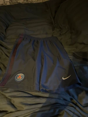 Blå PSG x Nike Dri-Fit shorts - Mörkblå PSG shorts från Nike med Dri-Fit material. Shortsen har elastisk midja, PSG-logga på ena benet och Nike swoosh på andra. Snygga röda detaljer längs sidan och sportig look. Perfekta för fotboll eller träning.