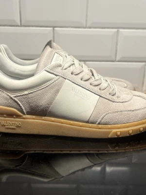 Valentino Garavani vita sneakers - Snygga sneakers från Valentino Garavani i vitt och beige mocka med detaljer i läder. Skorna har en klassisk låg siluett, vita skosnören och en beige gummisula. Perfekta för dig som gillar stilrena och exklusiva sneakers med premiumkänsla.