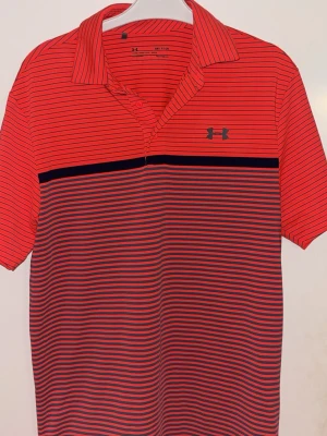 Röd randig pikétröja från Under Armour - Säljer en röd pikétröja från Under Armour med tunna svarta ränder och klassisk krage. Tröjan har korta ärmar och är tillverkad i ett lätt och bekvämt syntetmaterial. Perfekt för en sportig och avslappnad stil.