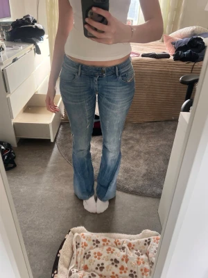 Bootcut jeans från Deisel - Säljer ett par snygga bootcut jeans från Diesel i ljusblå tvättad denim. Jeansen har klassisk femficksmodell, låg midja och lätt slitna detaljer för en avslappnad look. Perfekta till vardags med en enkel topp eller hoodie.