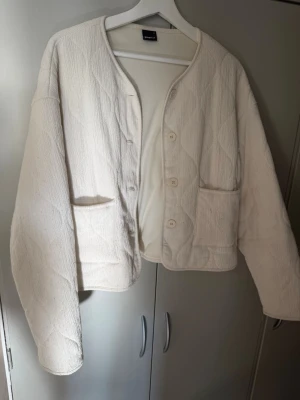Vit quiltad cardigan från Gina - Säljer en vit quiltad cardiganjacka från Gina med rund halsringning och knäppning framtill. Jackan har två stora fickor fram och är tillverkad i ett mjukt, strukturerat tyg. Perfekt för lager på lager och enkel att styla till olika outfits.