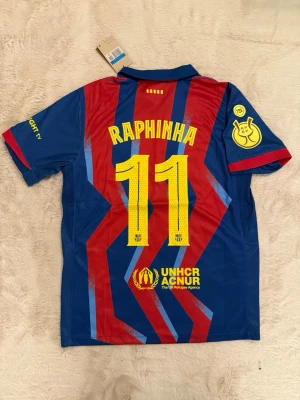 Barcelona matchtröja Raphinha #11 - Snygg Barcelona matchtröja från Nike med Raphinha och nummer 11 på ryggen. Tröjan är blå och röd med gula detaljer, klubbmärke och sponsorer som Spotify och UNHCR. Tillverkad i lätt och ventilerande material, perfekt för fotboll eller träning.
