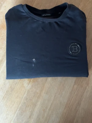 Svart t-shirt från Balmain - Snygg svart t-shirt från Balmain med rund halsringning och ett diskret B-emblem på bröstet. Tillverkad i mjuk bomull som känns skön mot huden. Perfekt för dig som gillar stilrena och exklusiva plagg. Lite smutsig på bilden men det går bort i tvätten 
