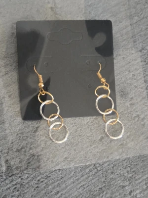 Hängande örhängen med ringar i guld och silver - Säljer ett par snygga örhängen med tre sammankopplade ringar i både guld- och silverfärg. Örhängena har krokar i guldfärg och en modern, minimalistisk design som passar till många olika stilar. Krokarna är i rostfritt stål och ringarna i legering. 