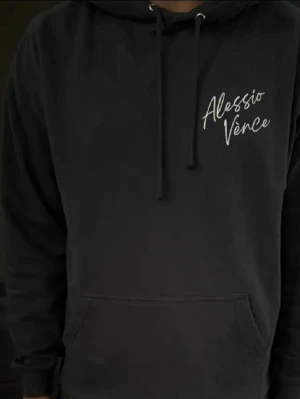 Svart hoodie från Alessio Vénce - Snygg svart hoodie från Alessio Vénce med vit logga på bröstet. Klassisk känguruficka framtill, justerbar dragsko i huvan och mjukt material som känns skönt mot huden. Perfekt för en chill och avslappnad stil. Inga defekter!
