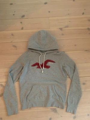 Grå hoodie från Hollister - Skit snygg hoodie från hollister med den klassiska fågeln på framsidan. Vit snörning vid luvan. Slimfit passform. Det står på etiketten att den är i storlek L, men det stämmer inte. Den passar S, jag är 186 och den är för liten på längden. 