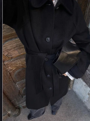 Svart ullkappa med knytskärp helt ny med prislapp  - Stilren svart kappa i ullmix med klassisk krage, knäppning framtill och matchande knytskärp i midjan. Kappan har raka ärmar och diskreta fickor, perfekt för en trendig och tidlös look. Nypris:1599kr