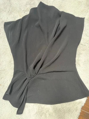 Svart omlottopp från Zara - Stilren svart omlottopp från Zara med knyt i sidan och asymmetrisk design. Toppen har en elegant drapering och är tillverkad i ett mjukt, lätt material som ger en snygg passform. Perfekt för dig som gillar minimalistisk och trendig stil.